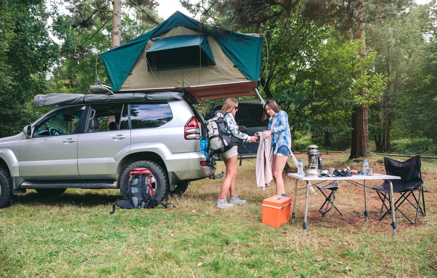 Zwei Frauen beim Aufbau eines Zeltlagers im Wald neben einem silbernen SUV, der mit einem Dachzelt und einer Markise ausgestattet ist; daneben stehen ein Campingtisch und Stühle.