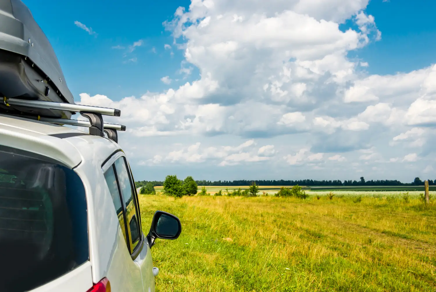 Nahaufnahme eines weißen Autos mit Dachgepäckträger und Dachbox, das am Rand einer saftigen grünen Wiese mit Blick auf eine ländliche Felder- und Wolkenlandschaft steht.