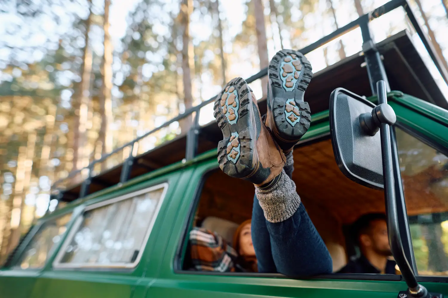 Eine Person streckt die Beine in Wanderschuhen durch das geöffnete Fenster eines grünen Campingbusses nach draußen, während sie im Wald entspannt.
